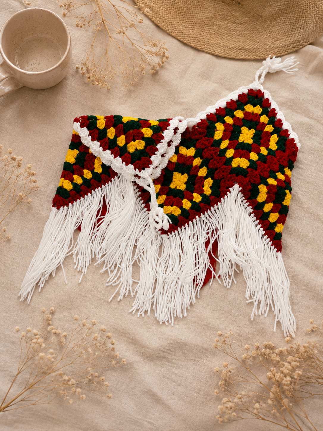 Folded Boho Chic Crochet Halter Top in red, green, yellow and white with long fringe, styled on beige linen with dried flowers and a ceramic mug.