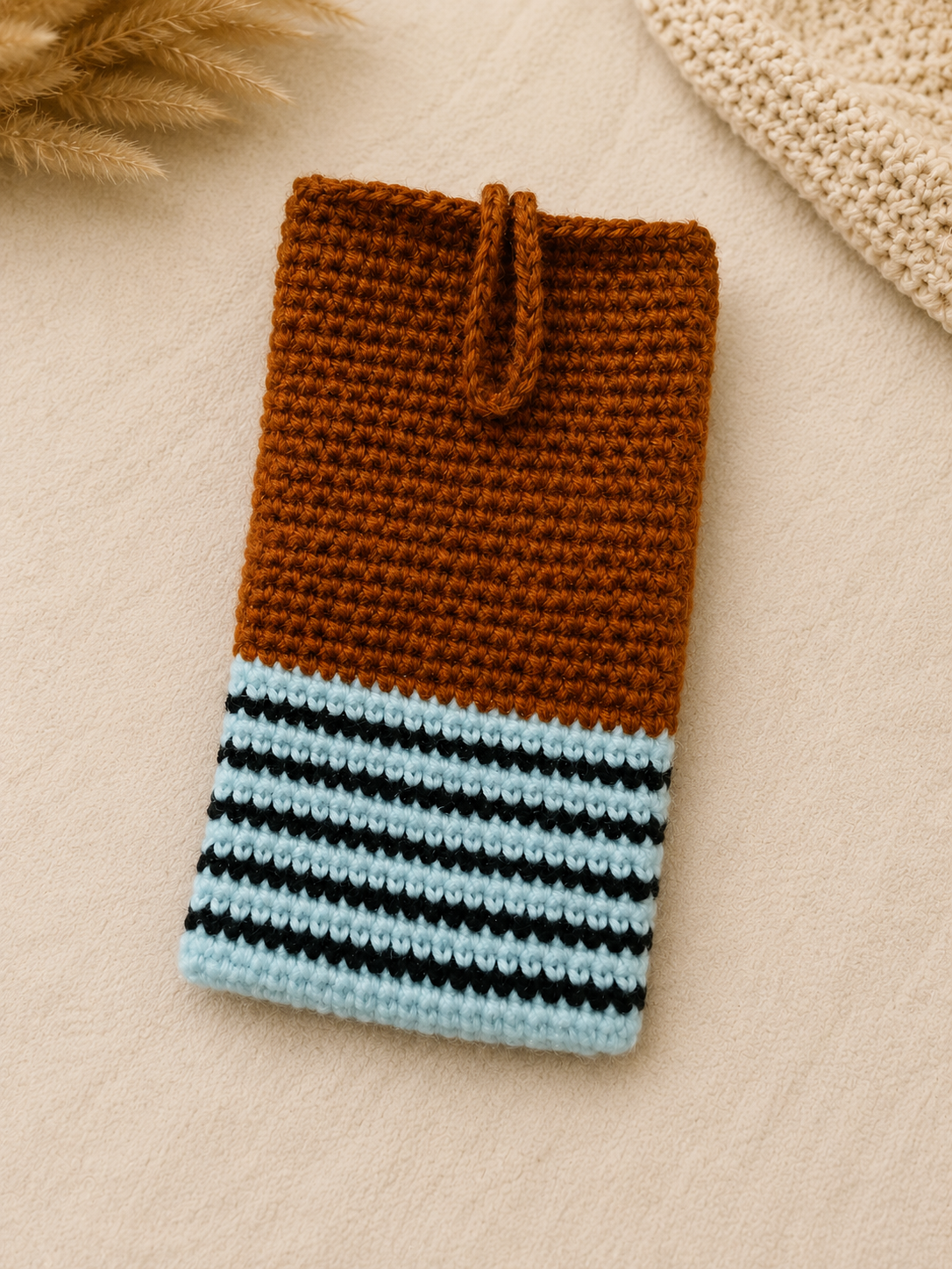 Rustic Loops crochet phone pouch in brown, light blue, and black stripes, displayed on a soft neutral background