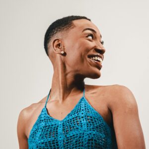 A woman wearing a blue crochet top smiles while looking back in a studio setting with a white background.