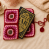 Pink and red handmade crochet book sleeve with floral granny square patterns, styled with a book, tea, candle, greenery, dried flowers, and soft cozy background props.