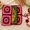 Pink and red handmade crochet book sleeve with floral square patterns holding a book, styled on a soft cream background with tea, candle, dried flowers, eucalyptus, and cozy knit accents.