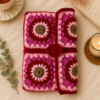Pink and red handmade crochet book sleeve with floral square patterns, styled on a warm cream background with tea, candle, dried flowers, eucalyptus, and cozy knit accents.