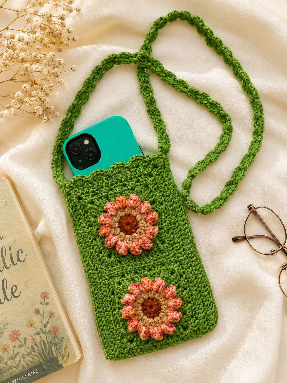 Side Sling Bag_1 Green handmade crochet side sling bag with two floral appliqués, shown with a phone inside on a soft cream fabric background.