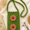 Side Sling Bag_2 Green handmade crochet side sling bag with two floral appliqués and a long strap, placed flat on cream fabric with lifestyle props.