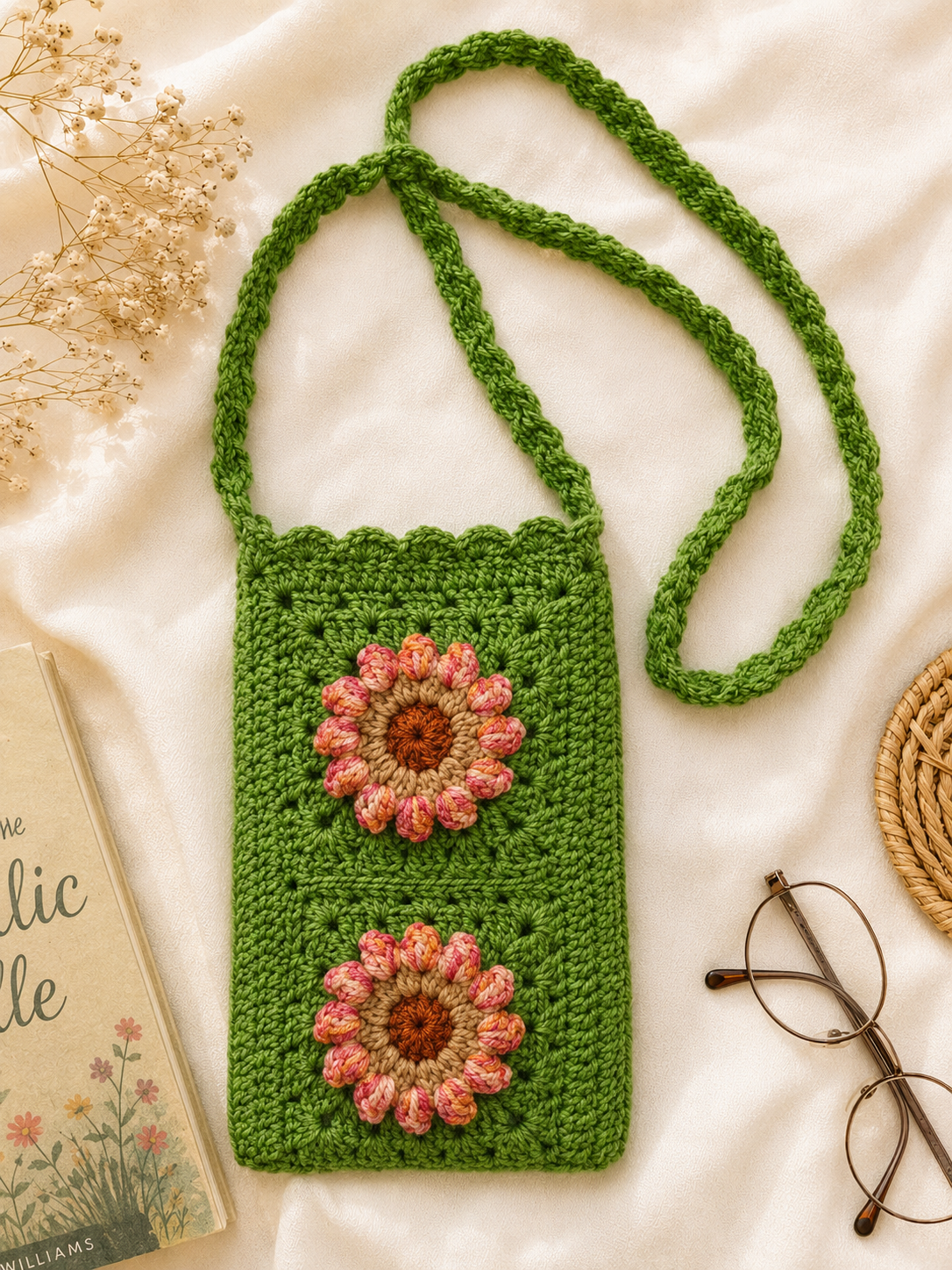 Side Sling Bag_2 Green handmade crochet side sling bag with two floral appliqués and a long strap, placed flat on cream fabric with lifestyle props.