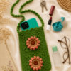 Green Crochet Side Sling Bag with Everyday Essentials Green handmade crochet side sling bag with floral appliqués, shown with a phone, keys, lipstick, earbuds, sanitizer bottle, eyeglasses, and a book on cream fabric.