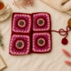 Handmade pink and red crochet book sleeve with floral granny square patterns, styled on a cream textured background with tea, candle, book, greenery, and soft decorative props.